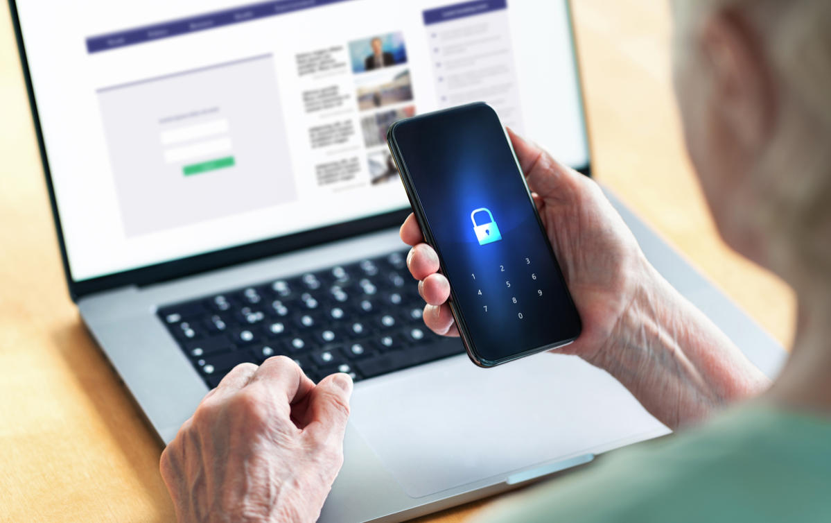 Older adult unlocking smartphone for secure login while seated at open laptop.