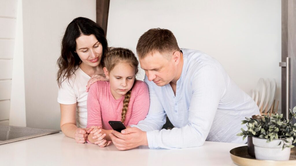 Parents guiding young daughter while viewing smartphone together at kitchen counter.